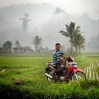 Far og sønn på en motorsykkel. I bakgrunnen er det en landsby og regnskog. Alt man kan se av regnskogen er dekket av røyk.